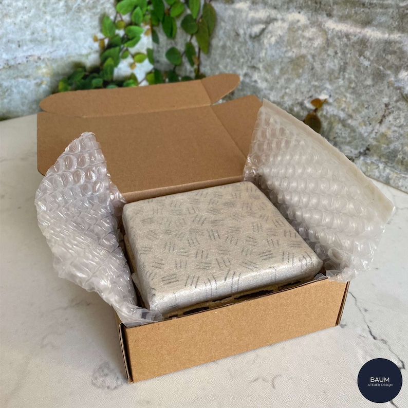 K&ouml;nnte beinhalten: Eine kleine, quadratische, graue und wei&szlig;e Keramikfliese mit geometrischem Muster ist in einem braunen Karton mit Luftpolsterfolie zum Schutz verpackt. Der Karton ist mit dem Text "BAUM ATELIER DESIGN" beschriftet.