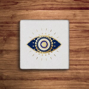 K&ouml;nnte beinhalten: Ein wei&szlig;er quadratischer Untersetzer mit einem blauen und goldenen b&ouml;sen Auge. Das Auge ist von einem goldenen geometrischen Muster und Lichtstrahlen umgeben.