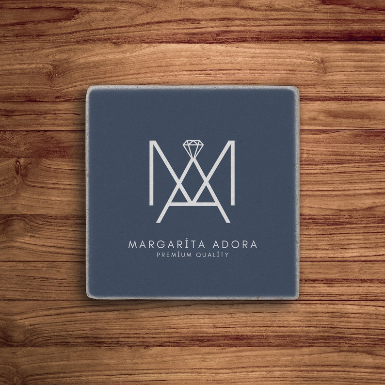 May include: A blue square coaster with a white logo. The logo is a stylized "M" with a diamond in the center. The text "MARGARITA ADORA PREMIUM QUALITY" is below the logo.