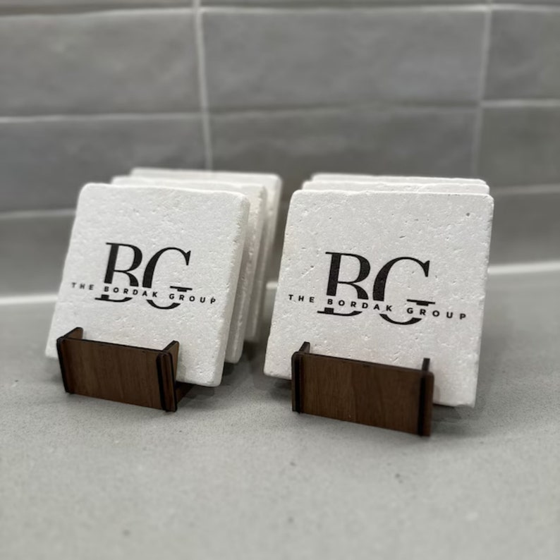 Puede incluir: Un juego de posavasos blancos con el texto "The Bordak Group" y las iniciales "BG" en negro. Los posavasos se muestran en un soporte de madera.