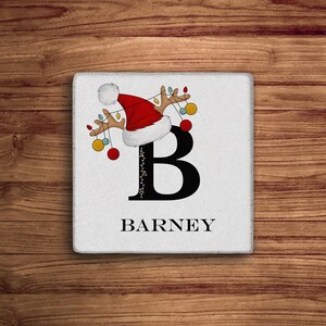 May include: White ceramic coaster with a black letter B, a red Santa hat with white trim, and reindeer antlers with Christmas lights and ornaments. The name "BARNEY" is printed below the letter.