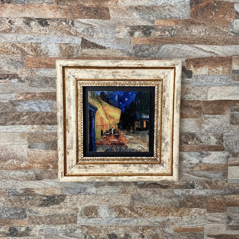 May include: A framed painting of a cafe at night with a starry sky. The painting is in a white frame with gold accents. The cafe is lit up with yellow lights and there are people sitting at tables outside.