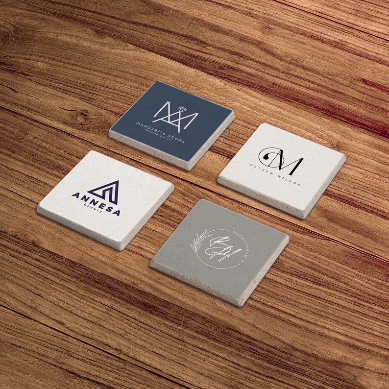 May include: Four square coasters with different designs. The coasters are made of a light gray material with a rough texture. The designs are all in black and white and feature initials and floral elements. The coasters are arranged in a square shape on a wooden surface.