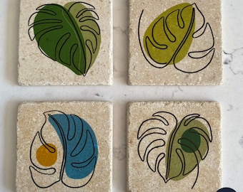 Op de afbeelding: Set van vier vierkante onderzetters met een beige achtergrond en een zwarte lijntekening van een monstera-blad. Elke onderzetter heeft een andere kleurvulling voor het blad, waaronder groen, geel, blauw en groen.