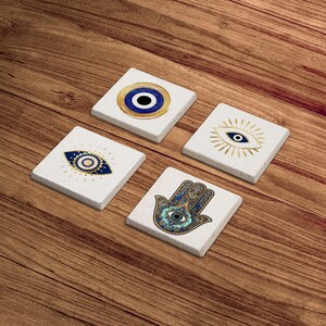 K&ouml;nnte beinhalten: Set aus vier wei&szlig;en Keramik-Untersetzern mit blauen und goldenen b&ouml;sen Augen-Designs. Ein Untersetzer zeigt eine Hamsa-Hand mit einem b&ouml;sen Auge in der Mitte.