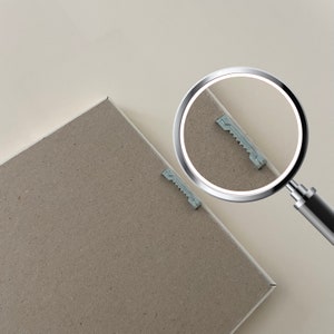 May include: A close-up of a brown cardboard backing with two small metal hanging clips. A magnifying glass is positioned over one of the clips, highlighting its details.