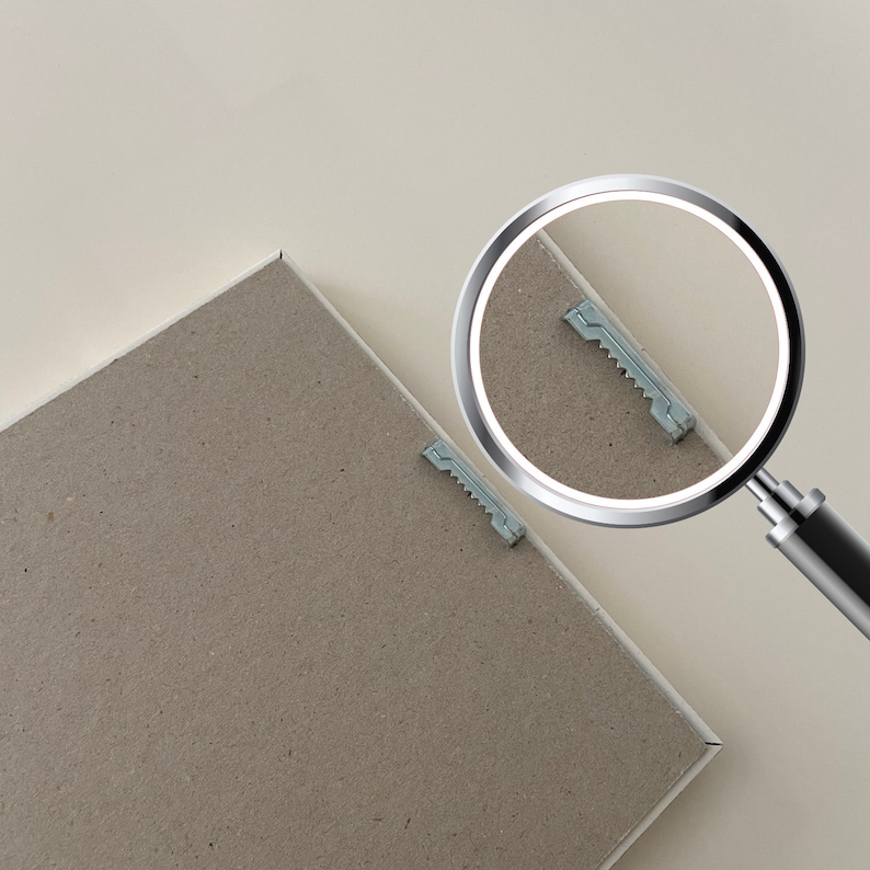 May include: A close-up of a brown cardboard backing with two small metal hanging clips. A magnifying glass is positioned over one of the clips, highlighting its details.