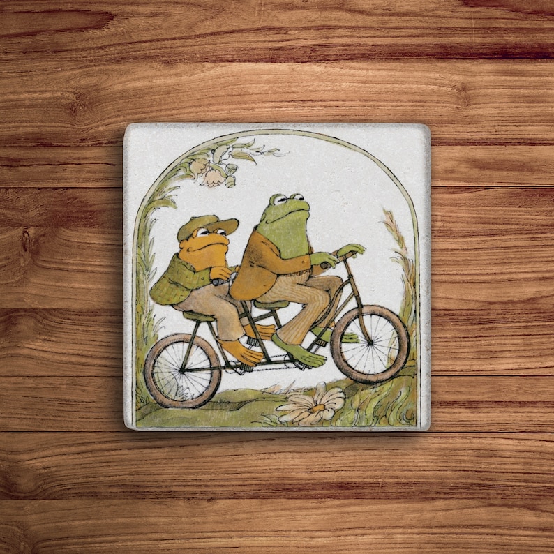 Puede incluir: Dos ranas montando una bicicleta t&aacute;ndem a trav&eacute;s de un paisaje verde y marr&oacute;n. La rana de delante es verde y la rana de atr&aacute;s es amarilla. La bicicleta tiene un cuadro blanco y ruedas marrones.