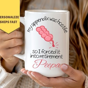 Peut inclure: Mug en céramique blanche avec le texte "my appendix was hostile so I forced it into retirement Peepa" et une illustration rose d'un appendice. Le mug est tenu par une personne.