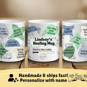 May include: Three white ceramic mugs with black outlines and green, blue, and gray illustrations. The mugs feature encouraging messages for someone recovering from an illness. The middle mug has the text "Lindsay's Healing Mug" and a progress bar that says "Recovery: Now Loading".