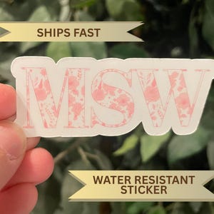 以下が含まれることがあります： ピンクの花柄の「MSW」の文字が入った白いステッカー。ステッカーは耐水性です。「SHIPS FAST」の文字がステッカーの上にあります。