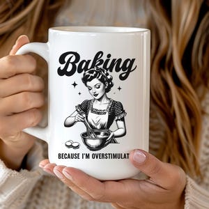 Peut inclure: Une tasse en céramique blanche avec une illustration en noir et blanc d'une femme en train de faire de la pâtisserie. Le texte "Baking because I'm overstimulated" est imprimé sur la tasse.