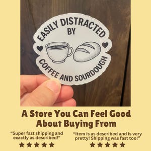 May include: A white sticker with the text "EASILY DISTRACTED BY COFFEE AND SOURDOUGH" in a circular design. The design includes a coffee cup, a loaf of bread, and two hearts. The sticker is held by a hand.