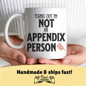 Peut inclure: Une tasse à café en céramique blanche avec du texte noir qui dit "Turns out I'm not an appendix person". La tasse a une illustration d'appendice rose. La tasse est tenue par les mains d'une personne.