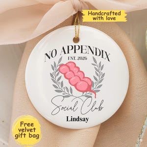 May include: A round white ceramic ornament featuring a pink graphic of an appendix, grey laurel leaf accents, and black text that reads NO APPENDIX EST. 2026 Social Club Lindsay. The ornament includes a gold hanging string at the top.
