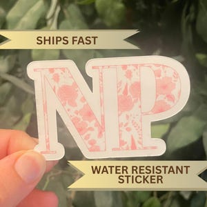 May include: A white sticker with the letters "NP" in a floral pattern of pink roses and leaves. The sticker has the text "SHIPS FAST" and "WATER RESISTANT STICKER" in a banner.