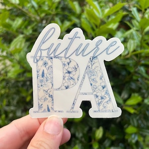 May include: A sticker with the word "future" in a cursive font above the letters "PA". The letters "PA" are filled with a blue floral pattern. The sticker is held in front of a green leafy background.