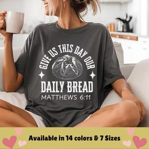 Op de afbeelding: Een donkergrijs T-shirt met witte tekst die "Give us this day our daily bread, Matthew 6:11" luidt. De tekst wordt omringd door een gestileerde broodvorm.