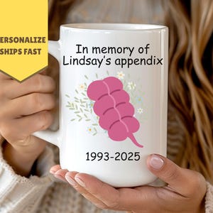 Peut inclure: Mug en céramique blanche avec l'inscription "In memory of Lindsay's appendix" et les années 1993-2025. La tasse présente une illustration rose d'un appendice avec des accents floraux. Une bannière jaune indique "Personalize Ships Fast".