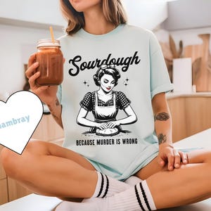 May include: Light blue t-shirt with the word "Sourdough" in a script font and a vintage illustration of a woman kneading dough. The text "BECAUSE MURDER IS WRONG" is below the illustration. A mason jar with a straw is held in the hand.