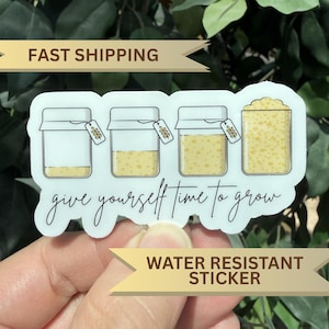May include: A white sticker with four jars, each with a different amount of yellow contents. The sticker has the cursive text "give yourself time to grow". It also has the text "WATER RESISTANT STICKER".