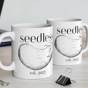 Könnte beinhalten: Zwei weiße Keramikbecher mit schwarzen Henkeln. Beide Becher zeigen eine Schwarzweißillustration einer Aubergine mit dem Text "seedless est. 2025" darauf.