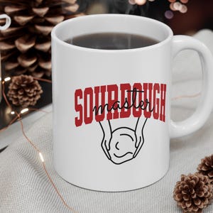 Peut inclure: Une tasse à café en céramique blanche avec un motif rouge et noir qui dit "Sourdough Master". La tasse a un contour noir de mains tenant une boule de pâte.