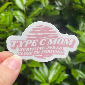 May include: A white sticker with pink text that reads "TYPE C MOM" above "SURVIVING AND SO CLOSE TO THRIVING". The sticker is held in front of a background of green leaves.