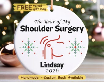 Può includere: Un ornamento rotondo in ceramica bianca con il testo nero The Year of My Shoulder Surgery, un grafico rosso di una spalla, due fiocchi di neve verdi e rossi e il testo nero Lindsay 2026. È appeso a un sottile cordino dorato.