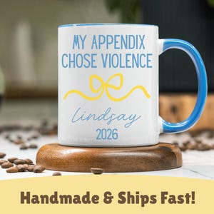 Peut inclure: Mug en céramique blanche avec une anse et un bord bleus, portant l'inscription "MY APPENDIX CHOSE VIOLENCE" en bleu, un nœud jaune, le nom "lindsay" et l'année "2026" en bleu. Le mug est posé sur un dessous de verre en bois.