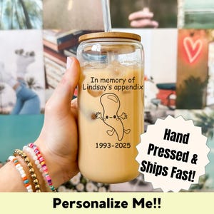 Puede incluir: Un vaso transparente con tapa de bambú y pajita, lleno de una bebida cremosa. El vaso está decorado con un diseño de apéndice de dibujos animados y el texto "In memory of Lindsay's appendix 1993-2025".