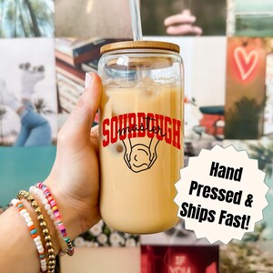 Puede incluir: Un vaso de vidrio transparente con tapa de bambú, lleno de una bebida de color claro y hielo, y una pajita. El vaso tiene las palabras "SOURDOUGH master" en letras rojas con un gráfico. Una pegatina dice "Hand Pressed & Ships Fast!"
