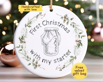 May include: A round white ceramic ornament featuring a black line drawing of a sourdough starter jar with the year 2026 printed on it. The text First Christmas with my starter is written in black, surrounded by a green botanical wreath of pine and holly.