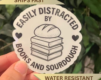 May include: A white, water-resistant sticker with the text "EASILY DISTRACTED BY BOOKS AND SOURDOUGH" in a circular design. The design includes a loaf of bread on top of a stack of books and two heart symbols. The sticker also says "WATER RESISTANT STICKER".