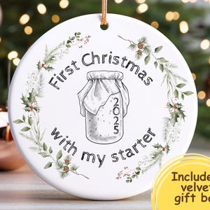 May include: White ceramic Christmas ornament with the text "First Christmas with my starter 2025". The ornament features a jar illustration and is decorated with a wreath of greenery and berries. Includes a velvet gift bag.
