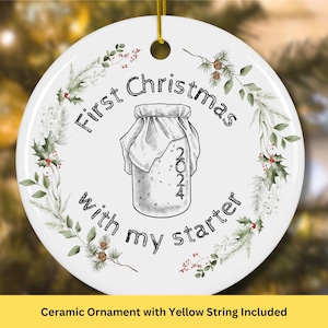 May include: White ceramic Christmas ornament with a green wreath design. The ornament features a hand-drawn illustration of a jar with a cloth lid and the text "First Christmas with my starter 2024".