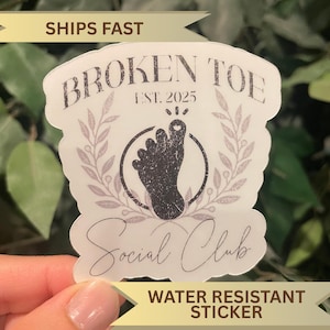 May include: A white, water-resistant sticker with the text "BROKEN TOE Social Club" and a graphic of a foot inside a circle. The sticker also includes the text "EST. 2025" and is decorated with a floral design.