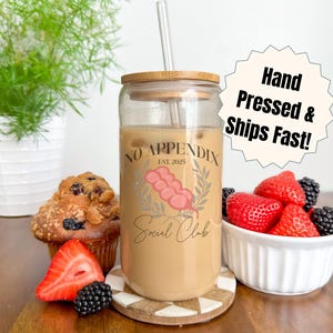 Peut inclure: Un verre transparent avec un couvercle et une paille en bambou, rempli d'une boisson brun clair. Le verre porte l'inscription "NO APPENDIX Social Club" et est accompagné d'un muffin, de fraises et de mûres.