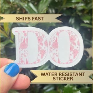 May include: A white sticker with the word "DO" in a pink floral design. The sticker is water resistant and has a gold banner that says "SHIPS FAST". The sticker is held by a hand with blue nail polish.