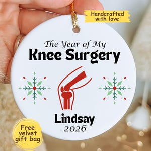 May include: A round white ceramic ornament featuring the black text The Year of My Knee Surgery, a red anatomical knee illustration, two green and red snowflakes, and the black text Lindsay 2026. A gold string is attached to the top for hanging.