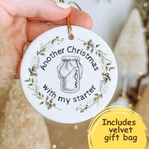 May include: A white ceramic Christmas ornament with the text "Another Christmas with my starter 2025" in a circular design. The ornament features a jar illustration and is decorated with a wreath of greenery. A gold string is attached for hanging.