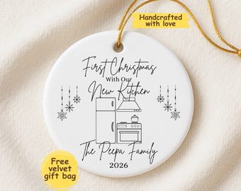 May include: A round white ceramic ornament featuring black line art of a refrigerator, stove, and range hood. The text reads First Christmas With Our New Kitchen, The Peepa Family, and 2026, accented by hanging snowflake illustrations and a gold string.