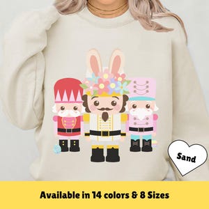 Puede incluir: Una sudadera beige con un gráfico de tres figuras de cascanueces. La figura central es un conejito con una corona de flores. El texto "Sand" está en un corazón blanco.