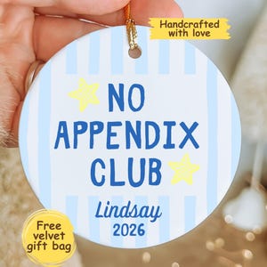 May include: A round white ceramic ornament featuring light blue vertical stripes and two yellow stars. The text reads NO APPENDIX CLUB, Lindsay, and 2026 in blue. A gold string is attached at the top for hanging, and a yellow tag says Handcrafted with love.