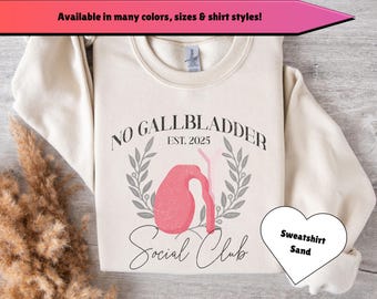 May include: A beige sweatshirt with the text "NO GALLBLADDER EST. 2025" and a pink organ graphic. The text "Social Club" is written below the graphic. The sweatshirt is available in sand color.