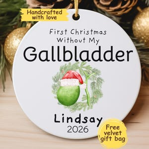 May include: White ceramic Christmas ornament with the text "First Christmas Without My Gallbladder." Features a green gallbladder with a Santa hat and the name "Lindsay" and the year "2026."