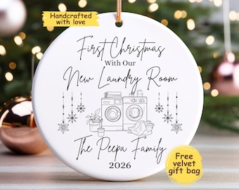 May include: A round white ceramic ornament featuring black line art of a washer and dryer, a potted plant, and hanging snowflakes. The text reads First Christmas With Our New Laundry Room, The Peepa Family, and 2026. It includes a gold hanging string.