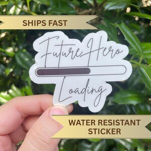 Könnte beinhalten: Ein weißer Aufkleber mit den Worten "Future Hero Loading" in handschriftlicher Schrift. Ein dunkelbrauner Ladebalken befindet sich in der Mitte. Der Aufkleber ist wasserfest.