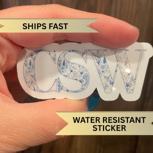 May include: A white sticker with the letters "CSW" in blue outlined letters. The letters are filled with a blue and white floral pattern. The sticker is water resistant.