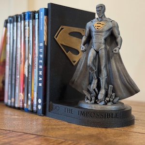 May include: A Superman-themed bookend set. The set includes a black bookend with a gold Superman logo and a gray Superman figure. The base of the figure reads "DO THE IMPOSSIBLE". Blu-ray discs are visible.